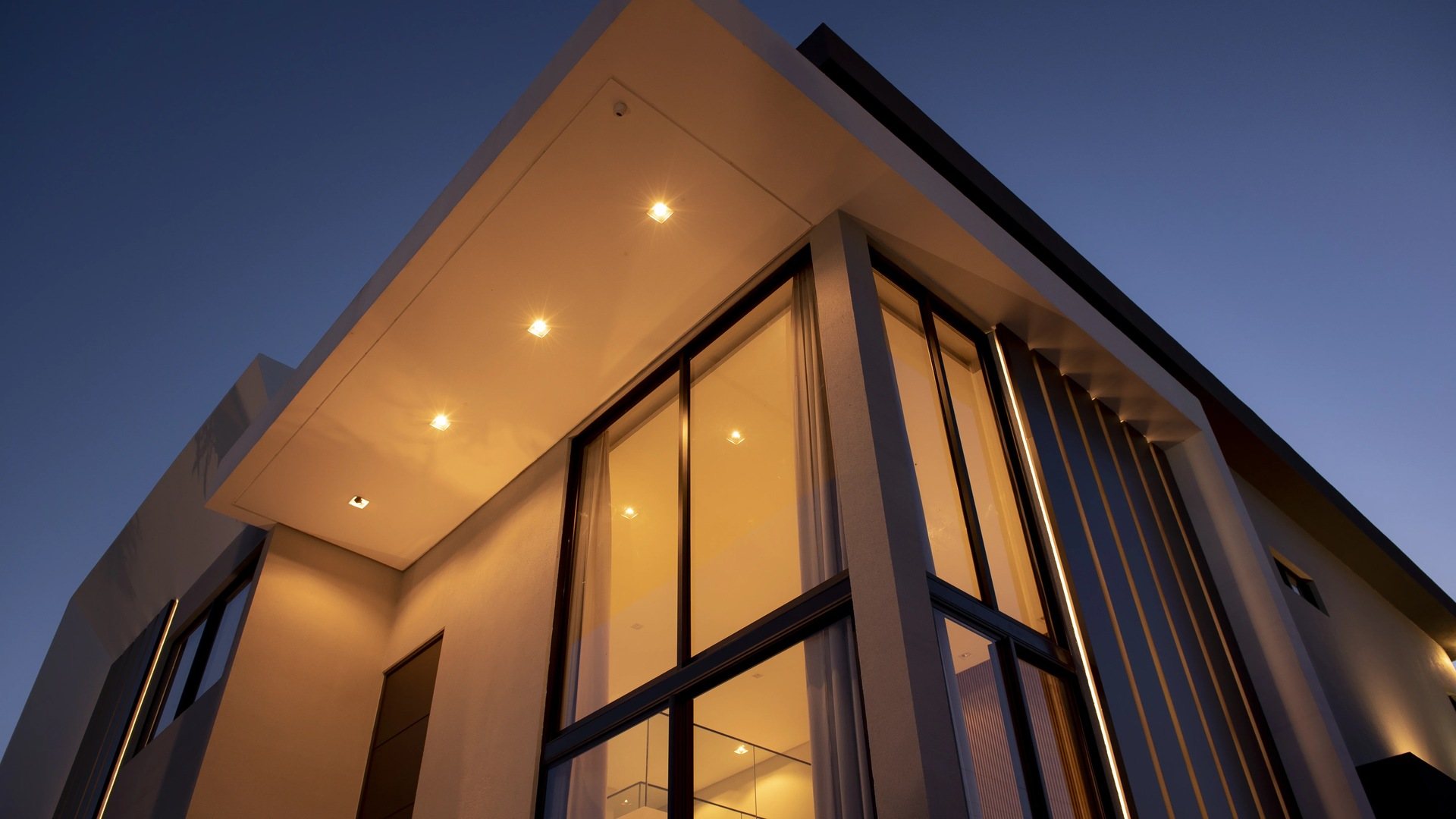 Casa GRAY ao entardecer: beiral branco em balanço com luzes acesas sobre a fachada envidraçada de pé-direito duplo.